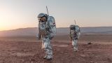 Two analog astronauts in silver Mars suits walk in line through the Negev Desert during daytime. There are many red stones and rocks on the ground and mountains in the background.